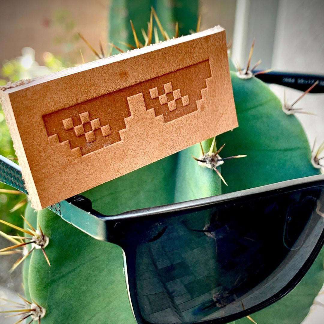 Veg-tan leather stamped with a pixel sunglasses design using a Delrin leather stamp, photographed resting on dark sunglasses outdoors.