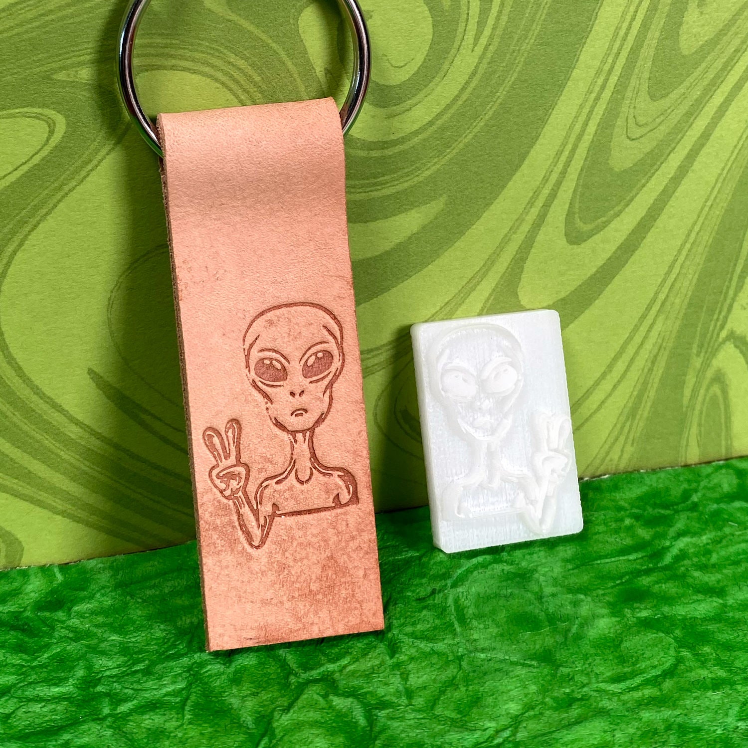 A rectangular leather keychain with an engraved alien holding up a peace sign is placed next to the Alien Peace Sign Delrin Leather Stamp, which features the same alien design. Both items are on a green textured surface with a green patterned background.