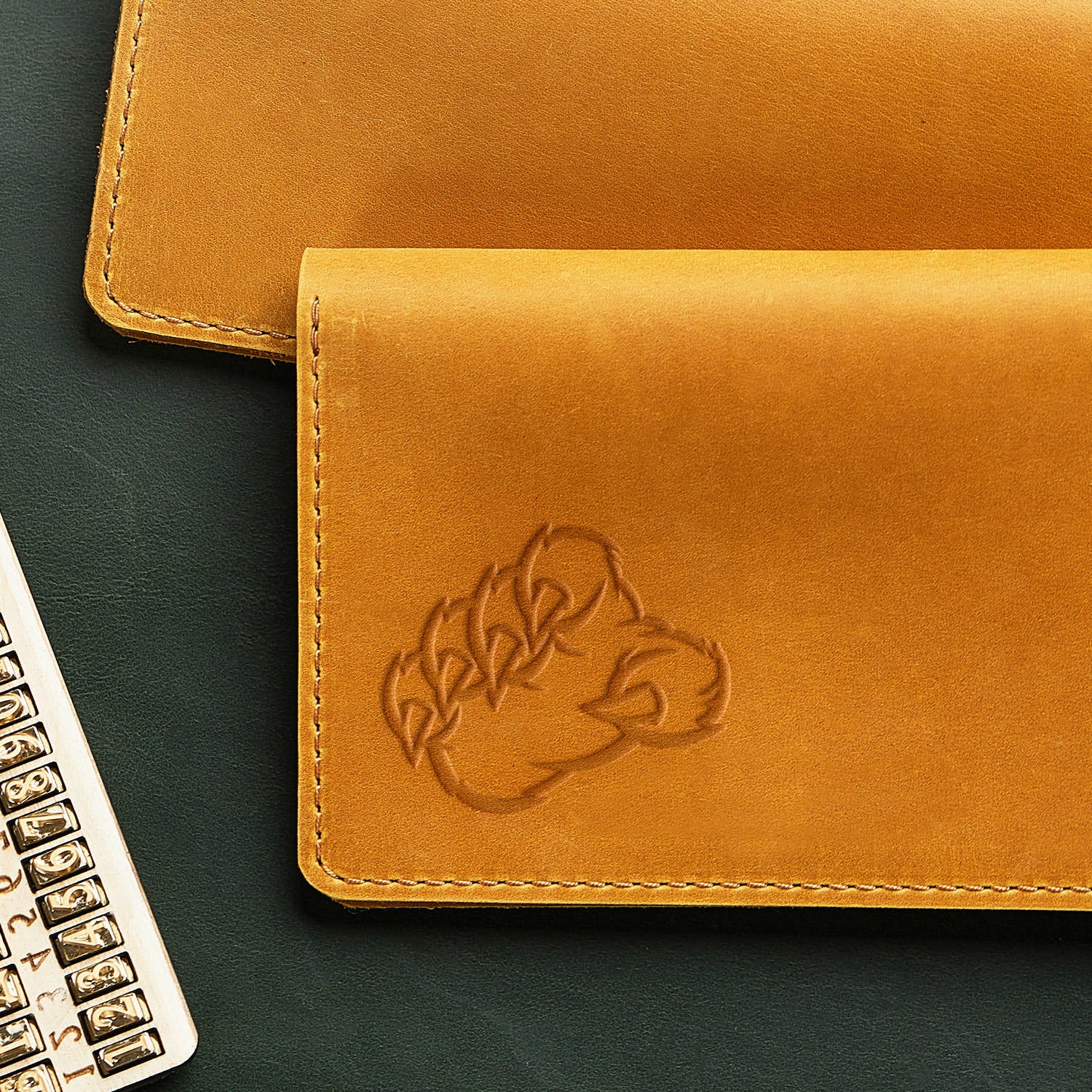 A Bear Claw Delrin Leather Stamp wallet is showcased on a dark green surface, exhibiting visible stitching around its edges. To the left of the image, there's a partially visible metallic keyboard with white keys.