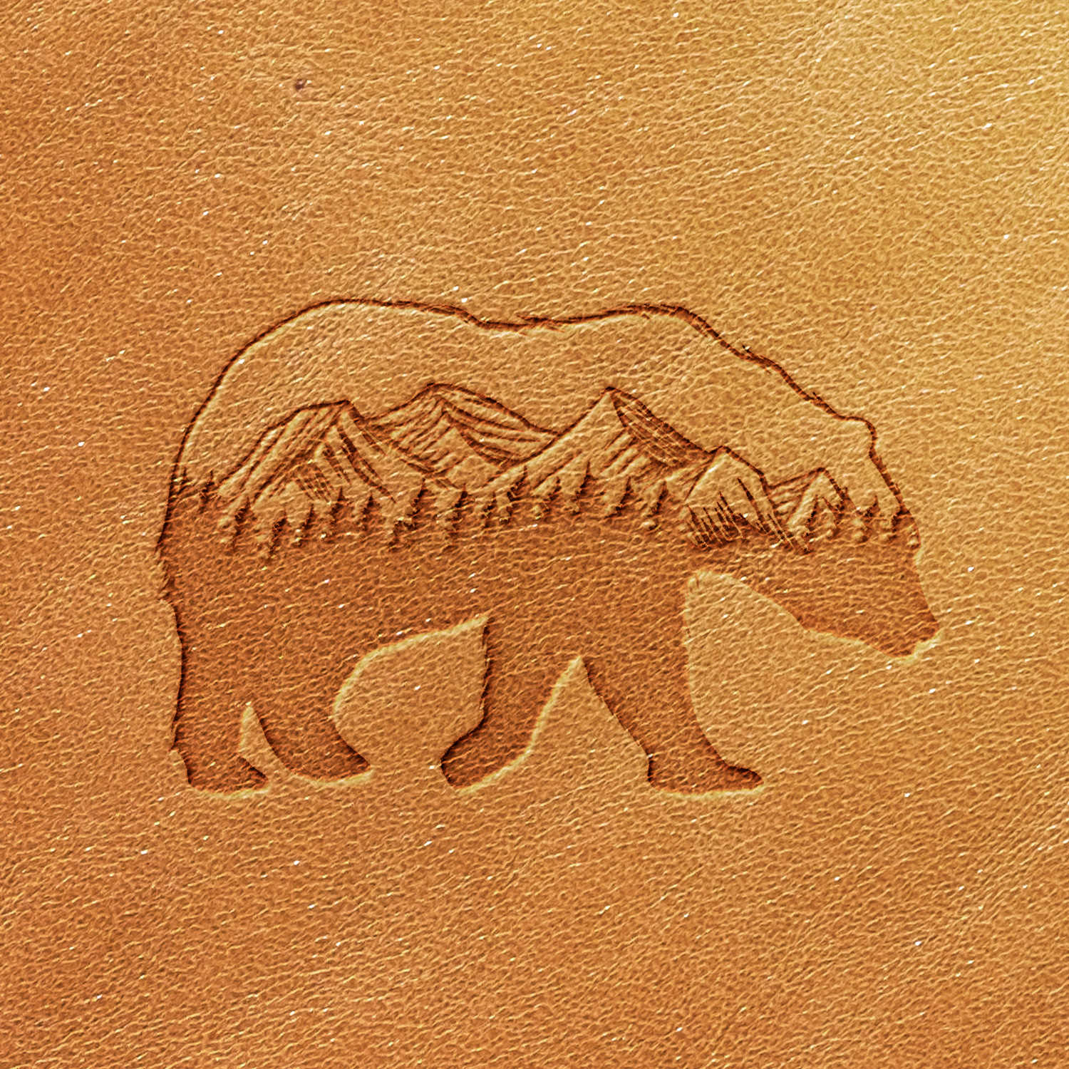 The Bear Mountain Scene Delrin Leather Stamp features a line drawing of a bear etched into a tan surface, with images of mountains and pine trees creating a nature scene within the bear's silhouette.