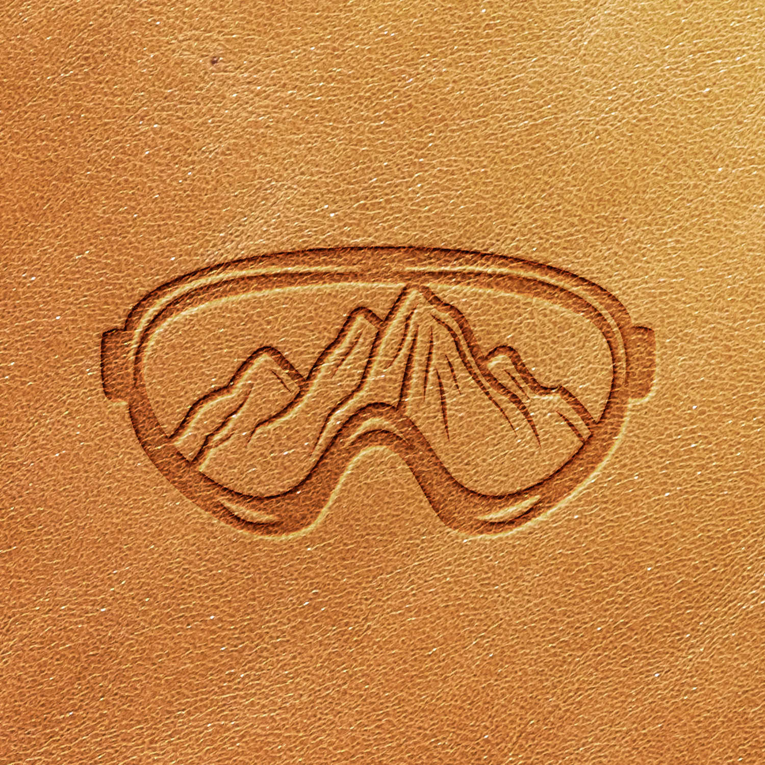 The Ski Goggles Delrin Leather Stamp creates a textured pattern on brown leather, featuring an embossed design of ski goggles encircling a mountain range.