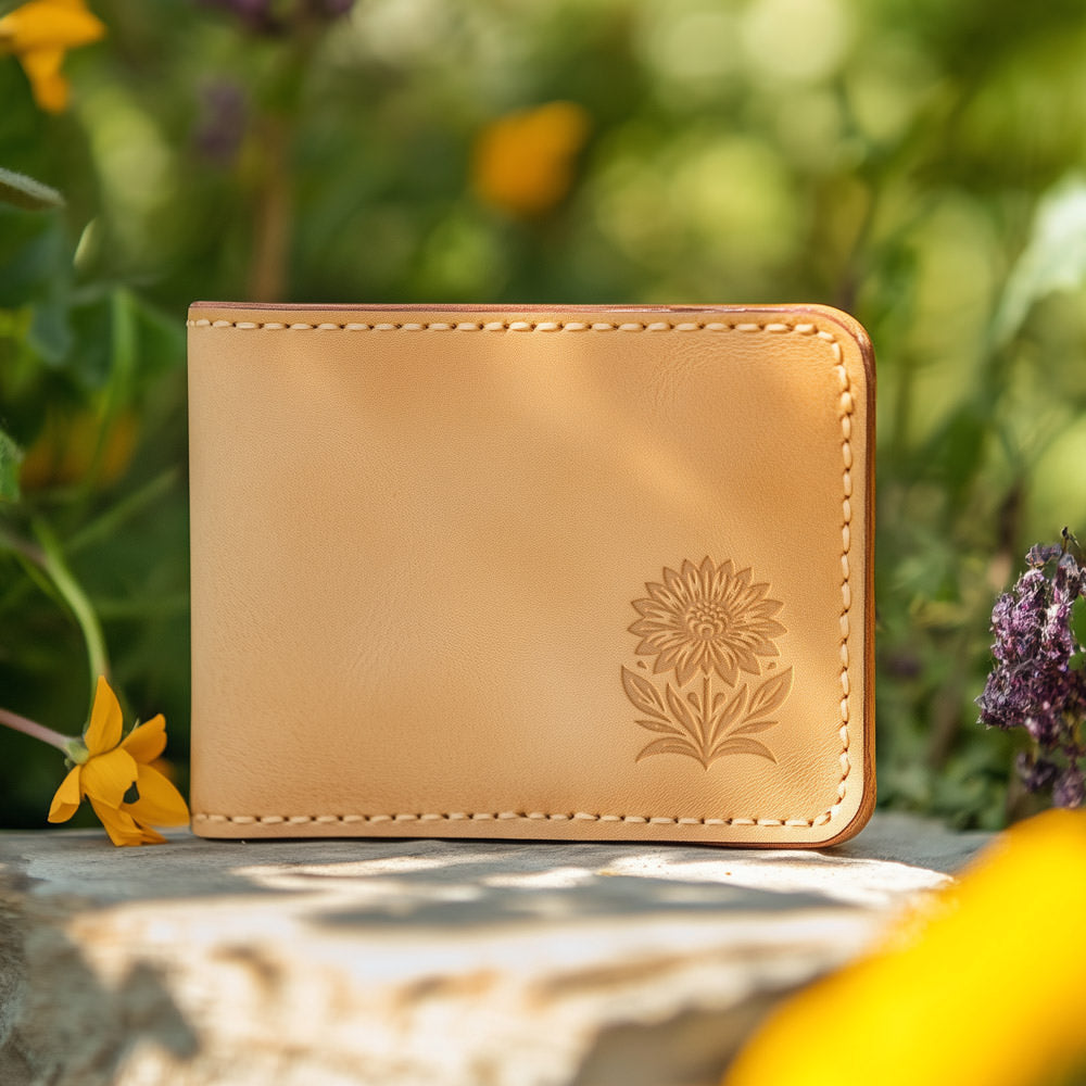 A light tan leather wallet featuring an embossed floral design created with the Sunflower Delrin Leather Stamp is placed outdoors. In the background, bright yellow and purple flowers along with green foliage are visible.