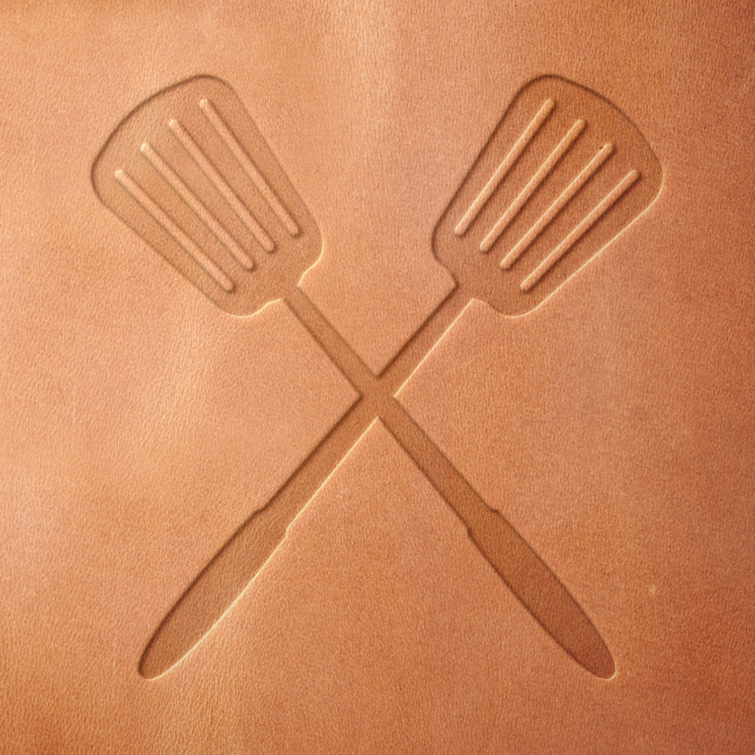 Leather sample showcasing a stamped crossed spatulas design using a delrin leather stamp, emphasizing the utensils’ parallel slots and balanced positioning.