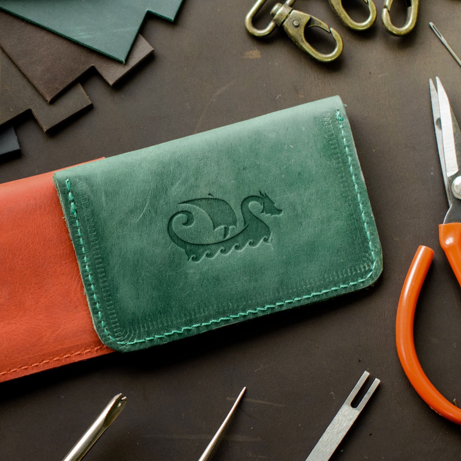 A green leather wallet sits on a brown surface with various leatherworking tools around it including scissors, a stitching chisel, a hole punch, two lobster clasps, and pieces of leather. The wallet has an embossed design of a Viking ship on the front flap from the Viking Ship Delrin Leather Stamp.