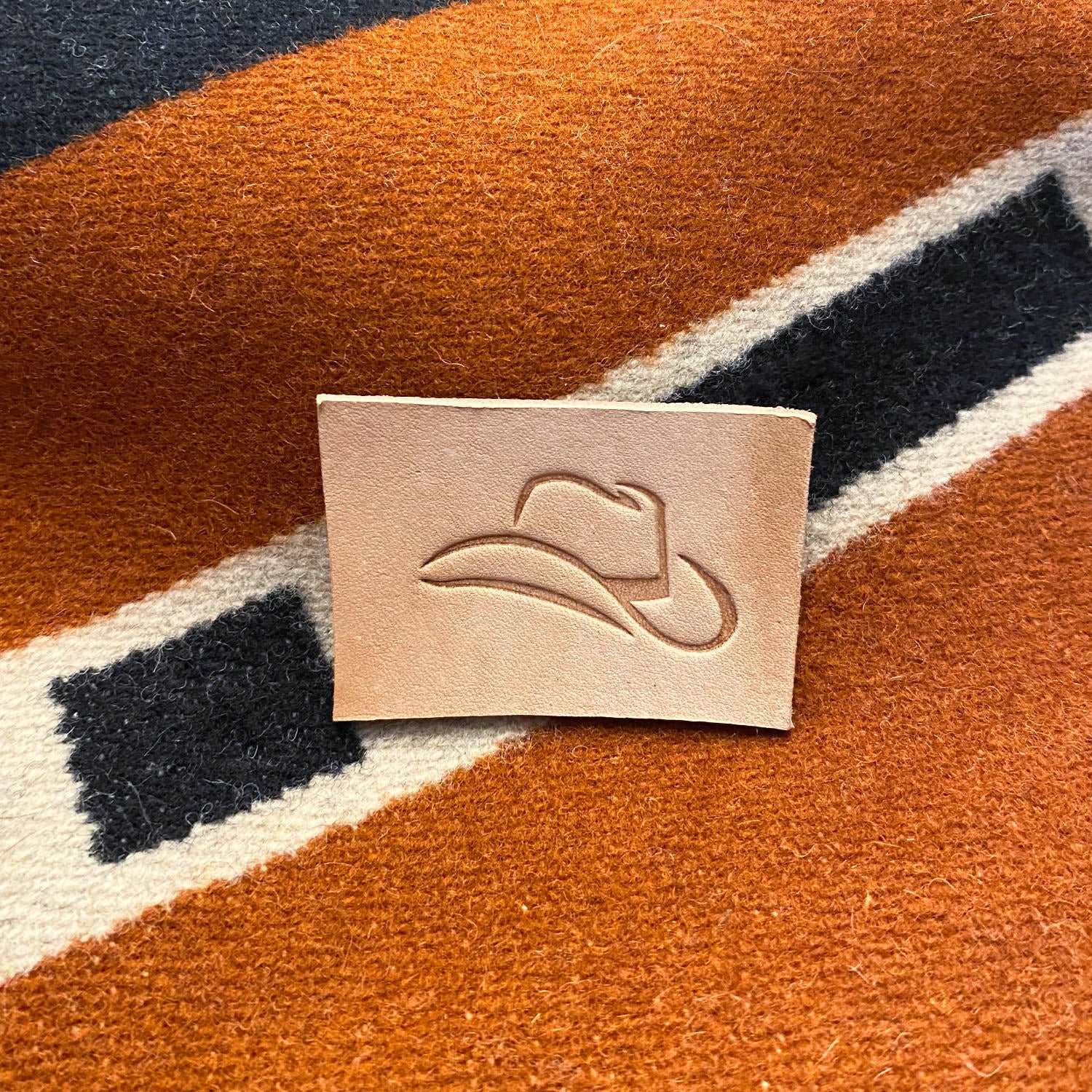 Small veg-tan leather sample stamped with a cowboy hat using a Delrin leather stamp, photographed on a woven western-style textile.