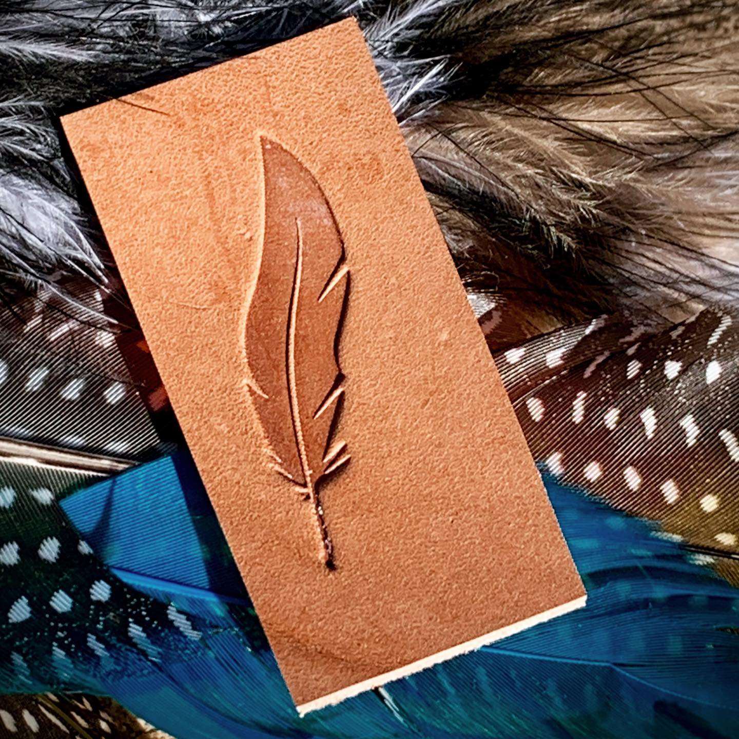 Natural veg-tan leather stamped with a feather design using a Delrin leather stamp, photographed on layered textiles with soft natural textures.