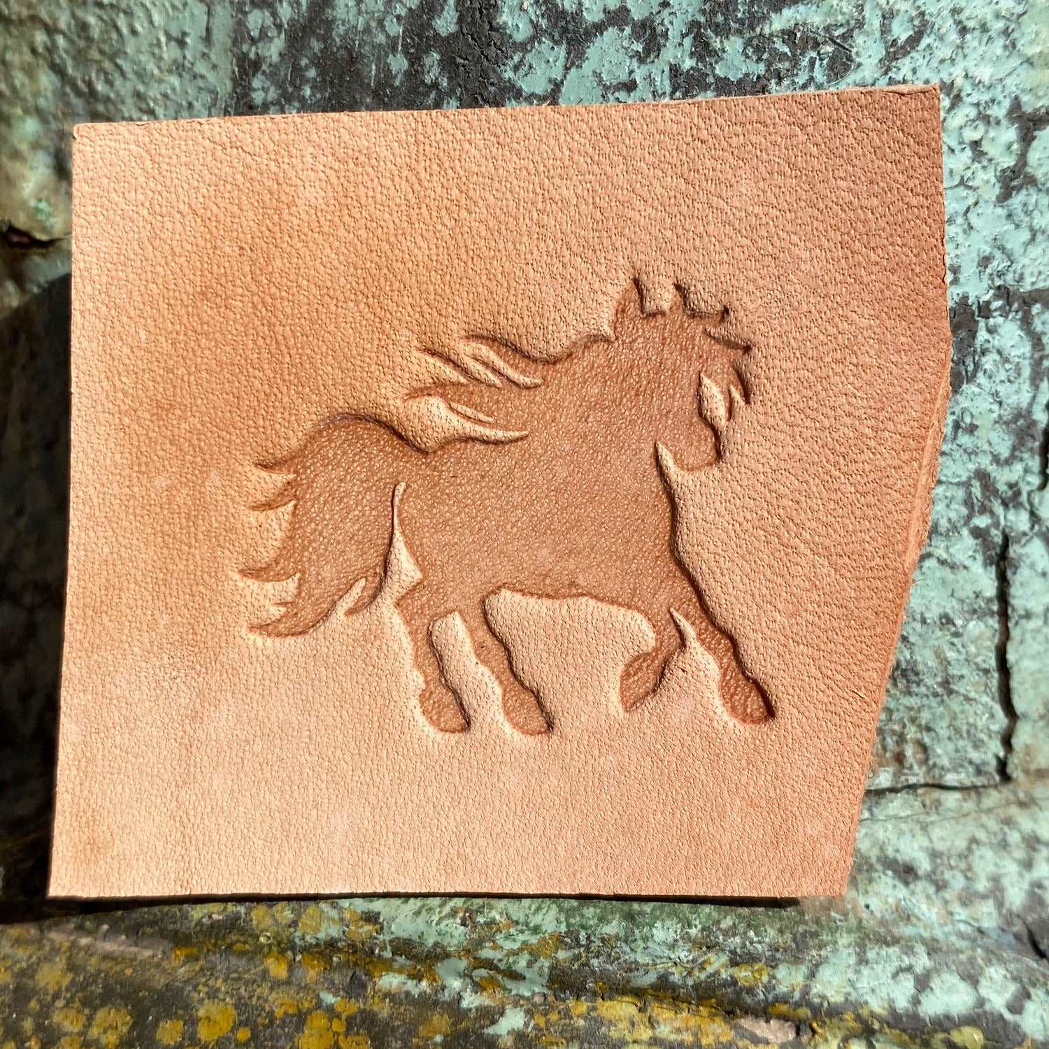 Natural veg-tan leather swatch stamped with a running horse silhouette using a Delrin leather stamp, photographed outdoors on stone.