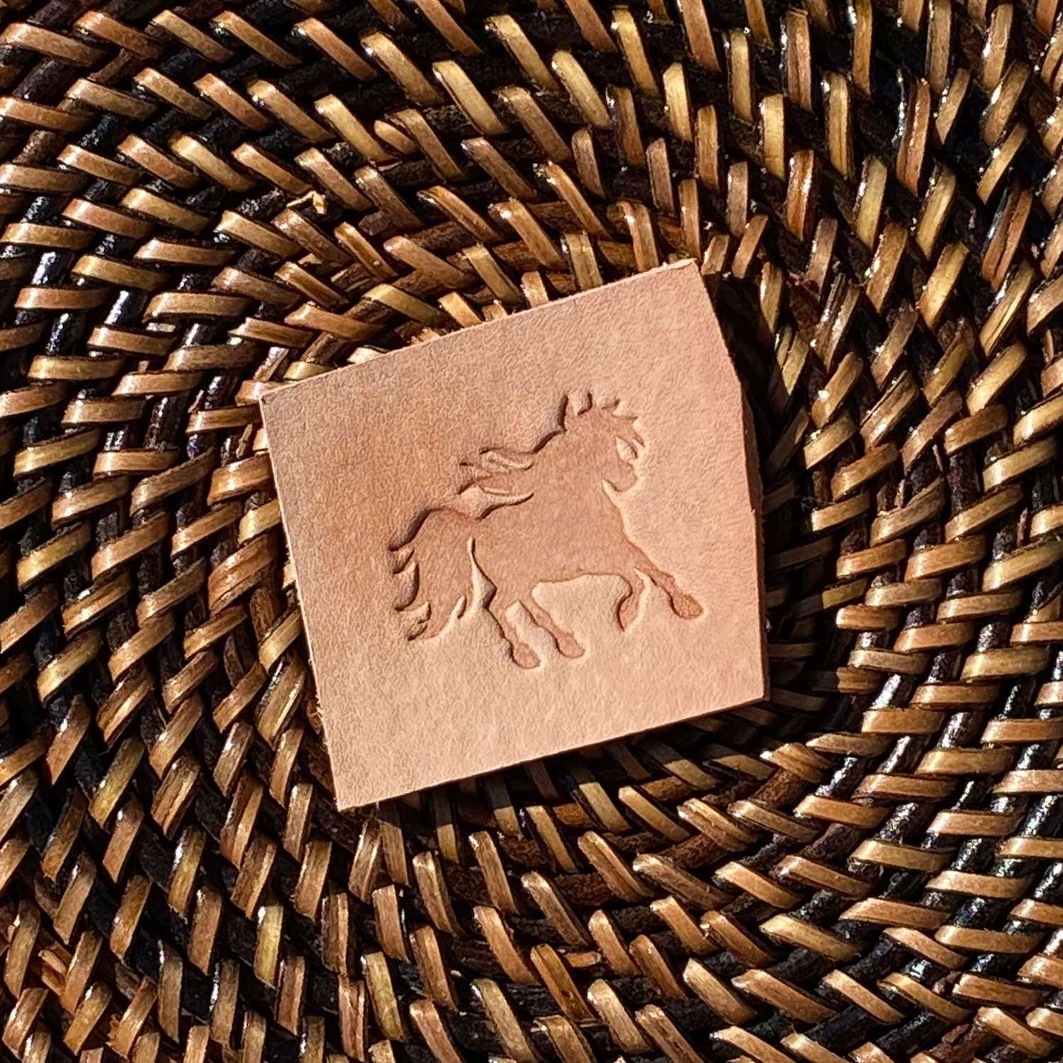 Small veg-tan leather sample stamped with a running horse silhouette using a Delrin leather stamp, styled on a woven surface.