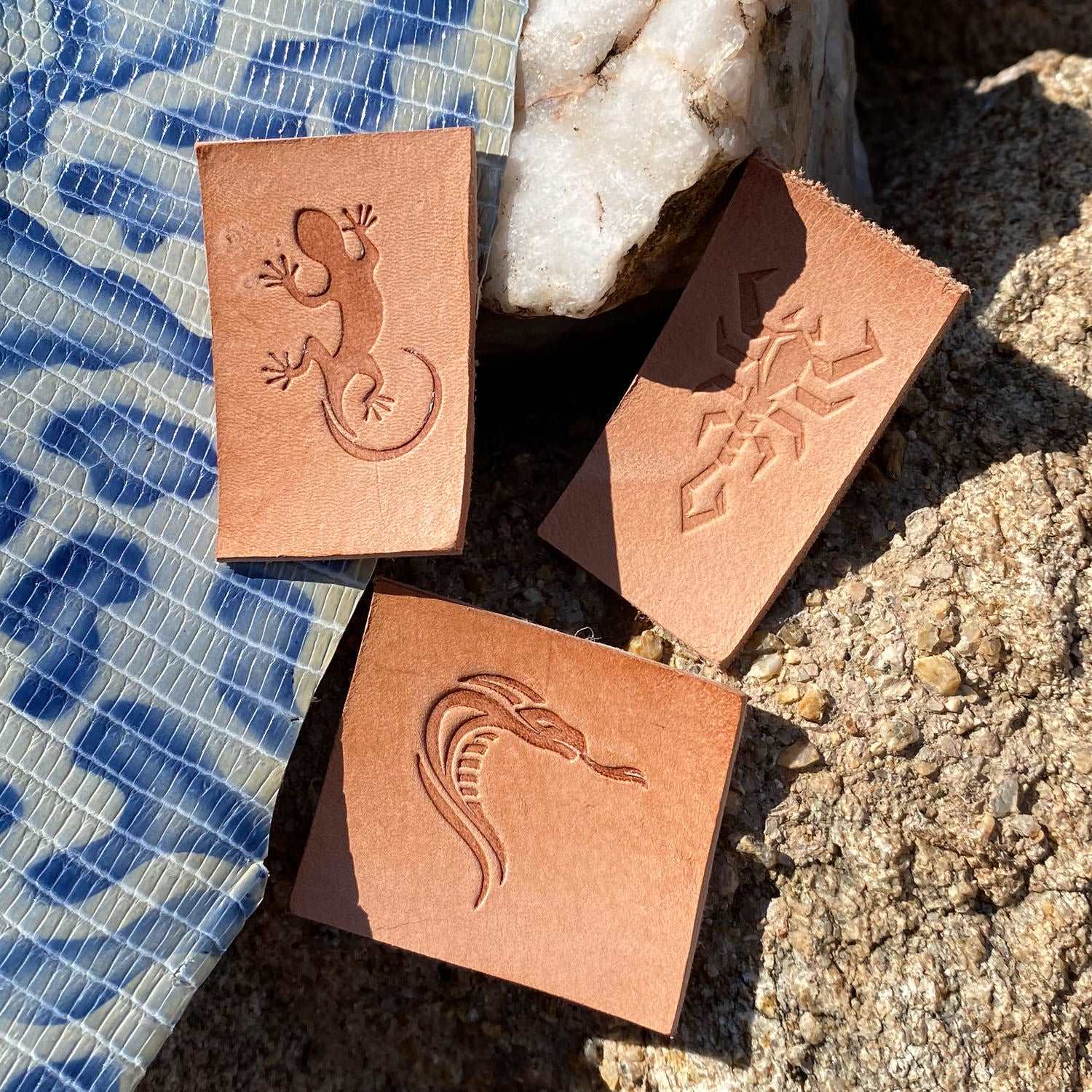 Assorted vegetable-tanned leather swatches stamped with snake designs using Delrin leather stamps, photographed outdoors on fabric and stone.
